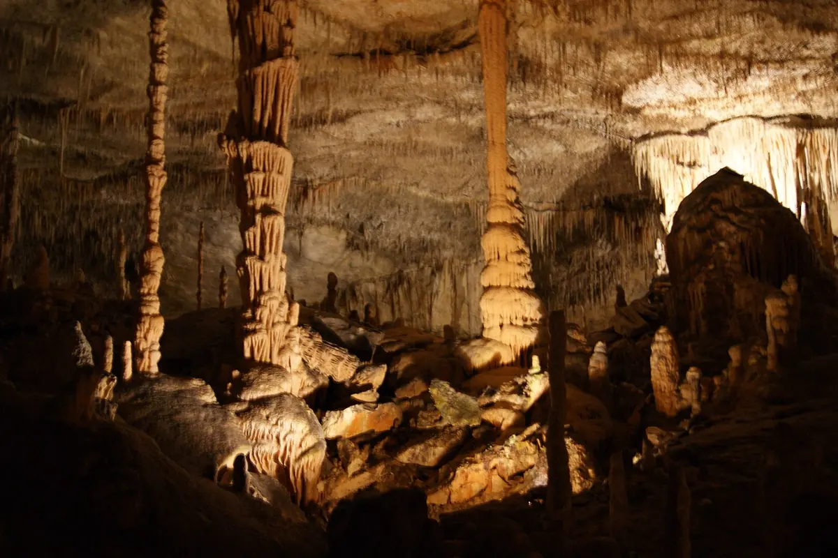 Tall pale stalagmite columns rising from a lit cave floor, with stalactites above and a dark chamber behind.